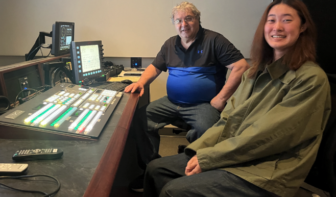 Two people smile at the camera. They are sitting in front of a recording booth