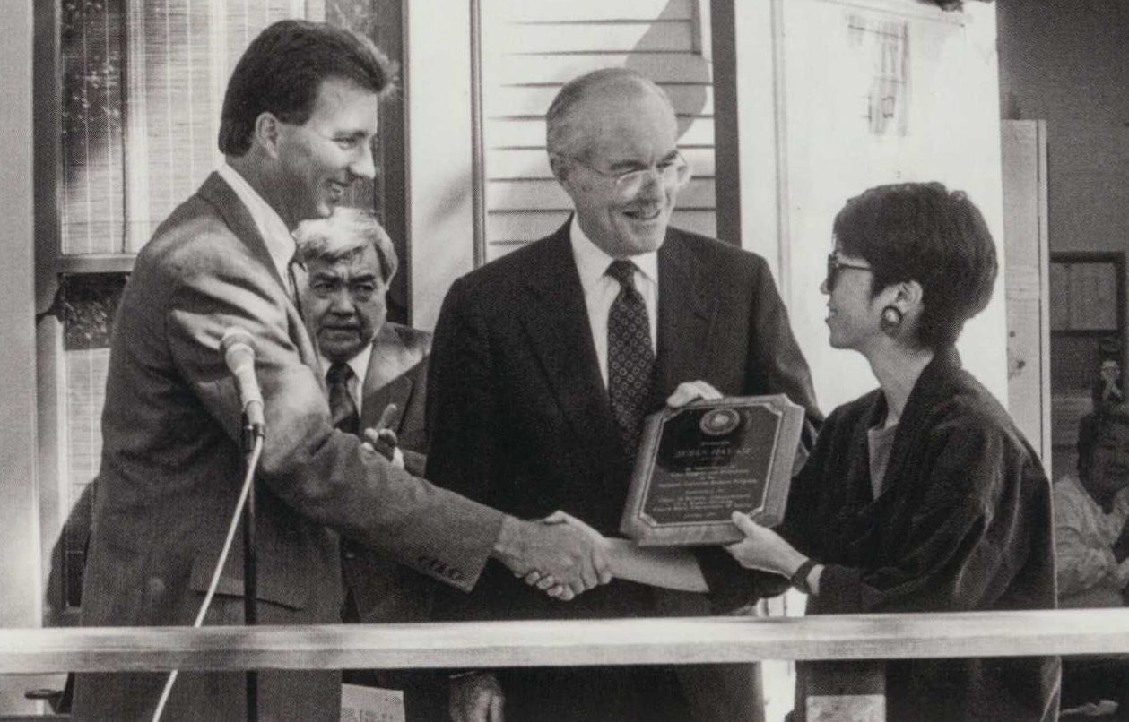 A black and white photo showing an Asian woman smiling and holding a plaque awarded to her from a White man.