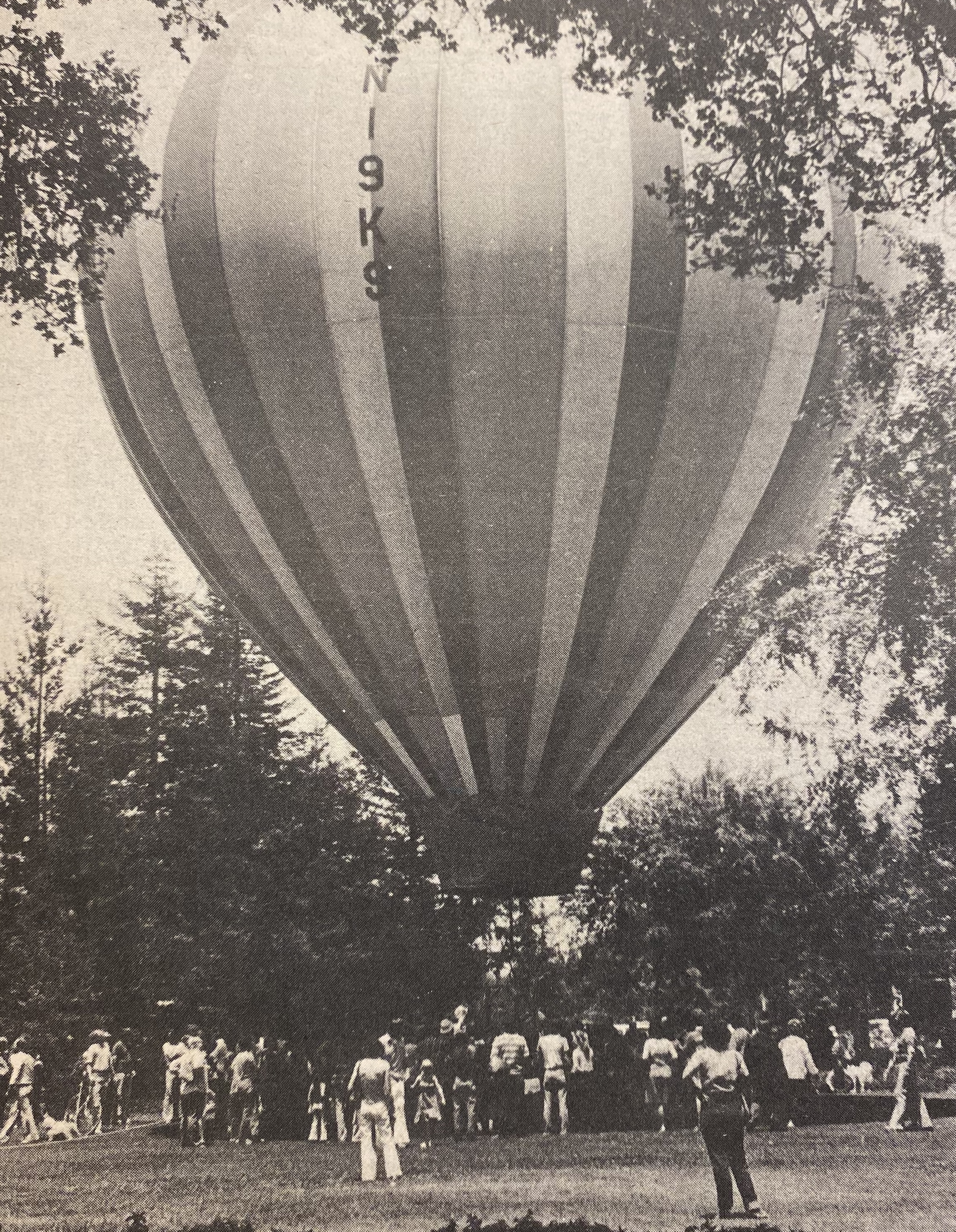 Black and white image of a hot air balloon with a number on it, surrounded by a group of people on a grassy field.
