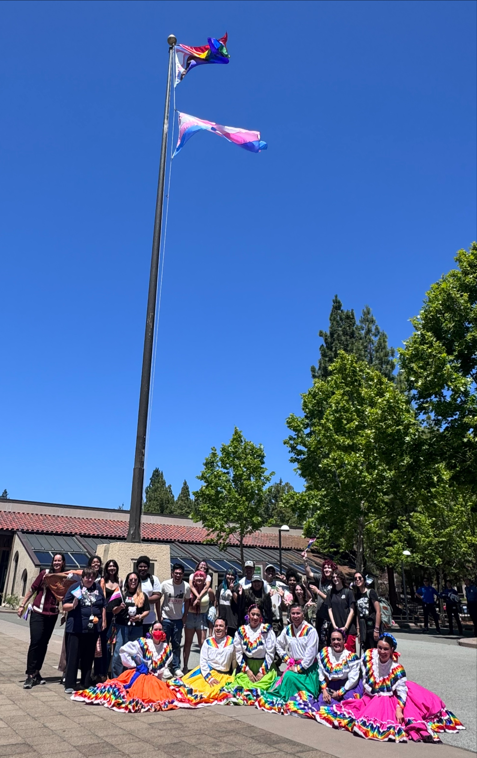 Pride flag-raising participants gather beneath the flags